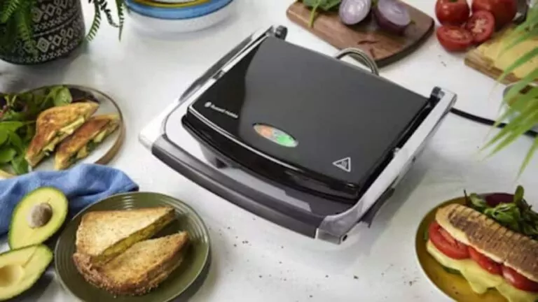 Sanduicheira multifuncional preta em uma mesa com sanduíches, abacate e ingredientes frescos ao redor, pronta para uso